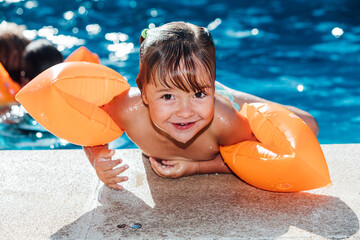 Little blond-haired girl in the pool wearing sleeves, smiling and looking at camera