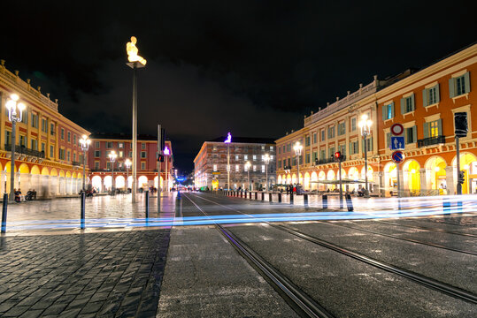 Car headlights make light trail from long exposure as the cross tram tracks late at night in Place Massena in the historic city of Nice, France.