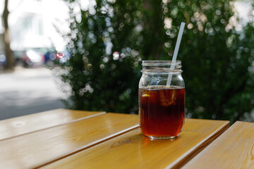 Close up outdoor view of a glass bottle of cold brew coffee with ice on wooden table and background of street.