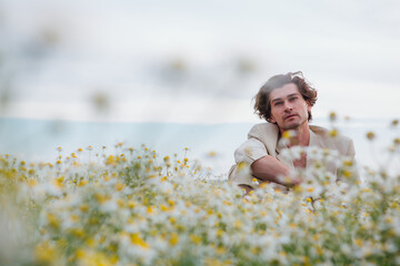 Fototapeta na wymiar Tall handsome man sitting on a white chair in camomile flowers field