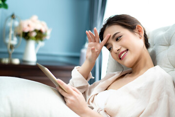 Beautiful charming Asian woman sitting on white sofa and using digital tablet connecting to social networking at living room in the morning. Happiness relaxation at home 