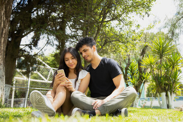 Hispanic young couple sitting in the park using smart phone on a sunny day - young people making video call with their phone