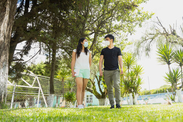 Young Hispanic couple walking with disposable face masks with social distancing - couple walking in a park on a sunny day