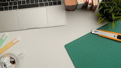 Workspace with copy space, laptop, cutting mat, stationery, books and utility knife on white desk