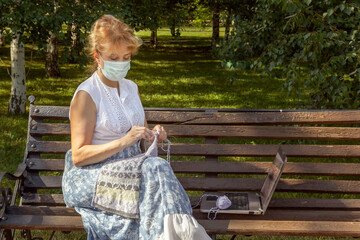woman with a medical mask on her face is sitting on a park bench knitting wool clothes on knitting needles and watching lessons or teaching knitting through a laptop. Distance learning. Copy space