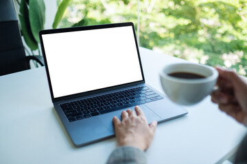 Fototapeta premium Mockup image of a businesswoman using and touching on laptop touchpad with blank white desktop screen while drinking coffee in office