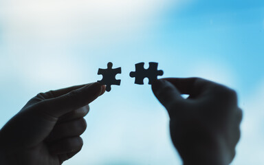 Closeup image of hands holding and putting a piece of white jigsaw puzzle together with blue sky...
