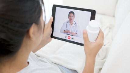 Telemedicine concept back view of asian woman video call with doctor at living room home. Patient girl consulting with general practitioner application computer tablet. Doctor and Consultant online