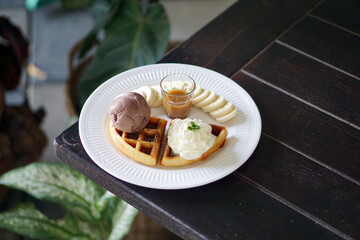 A plate of Belgian waffle with banana and caramel sauce, topped with whipped cream and chocolate ice cream.