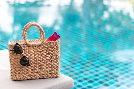 Weaving bag and hat with sunglasses and sunscreen lotion next to a swimming pool, summer travel concept