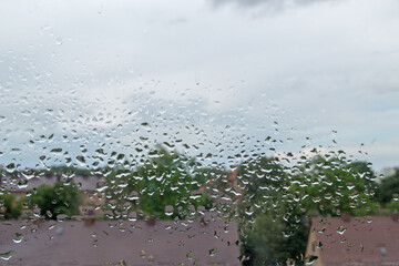 Chaotically arranged on the window glass raindrops make a amazing pattern against the background of gray blue cloudy sky and blurred brown roofs of low buildings surrounded by greenery of trees below
