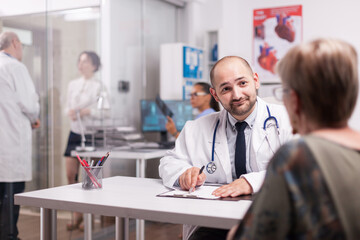 Young doctor and senior patient in hospital office discussing about illness treatment. Medic wearing white coat and stethoscope. Mature physician talking with sick woman in clinic corridor.
