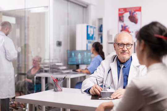 Senior Doctor Consulting Woman Patient In Hospital Office While Younger Colleague Is Discussing With Disabled Elderly Aged Woman In Wheelchair On Clinic Corridor And Nurse In Blue Uniform Looking At X