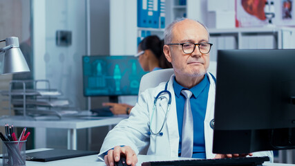 Naklejka na ściany i meble Elderly doctor working on PC in modern private clinic with glass walls. Hospital professionals in health industry staff at work in medicine medical practitioner specialist in unifor at desk job