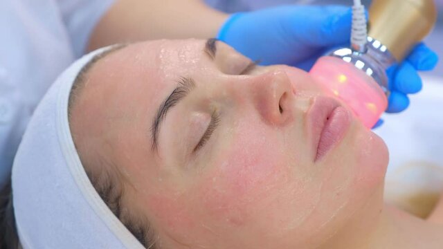Beautician cosmetologist doctor is moving manipula with hot red light on face. Portrait of woman on apparatus phonophoresis face procedure with hyaluronic acid gel in cosmetology clinic, side view.
