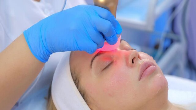 Cosmetologist doctor making phonophoresis procedure on woman face with hyaluronic acid gel, side view. Beautician moving manipula with hot red light on face. Apparatus procedure in cosmetology clinic.