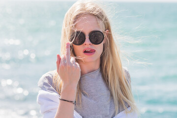 Provocative female hipster giving the middle finger fuck you to the camera at the beach wearing sunglasses. Offensive behavior hand gesture.