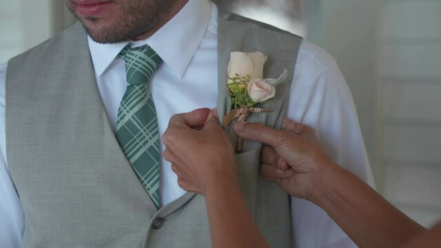 Caucasian Groom gets white rose Boutonniere pinned by woman on grey suit. Close up shot