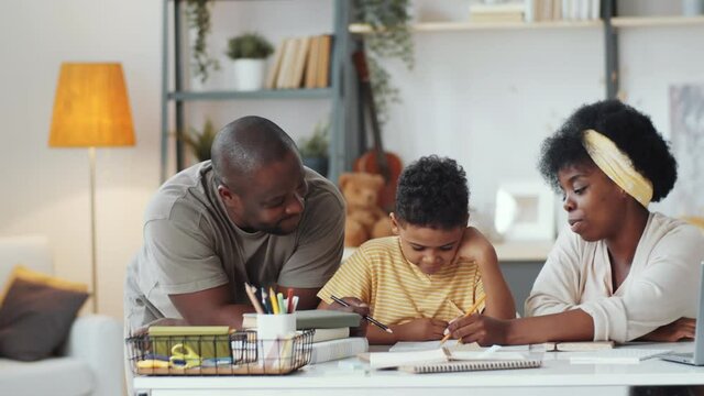 Joyous afro-american family doing homework together with little son, then looking at camera and smiling while sitting at table in the living room