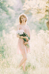 Fototapeta na wymiar Beautiful young Caucasian blond woman in light dress holding bouquet of white peonies, walking in summer field or garden in sunset. Woman with flowers outdoors