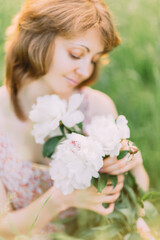 Beautiful young Caucasian blond woman in light dress holding bouquet of white peonies, walking in summer field or garden in sunset. Woman with flowers outdoors