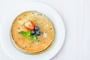 On a plate is a stack of hemp flour pancakes. Decorated with blueberries, strawberries, cannabis seeds and mint. Dusted with powdered sugar. Flat lay