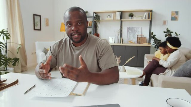 African American man looking at camera, showing line graphs on paper and discussing business plan on online video call while working remotely at home; wife and son resting on sofa in background