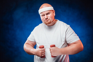 Corpulent funny man in a white tracksuit holds dumbbells in his hands and looks at the camera
