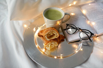 morning, hygge and breakfast concept - croissants, cup of matcha tea book and glasses in bed at home