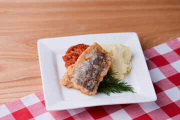 Fried fish and mashed potatoes with dill and gravy served on a white plate over light wooden table background.