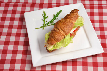 Breakfast with fresh croissants with meat and salad filling served on white plate over red plaid tablecloth.