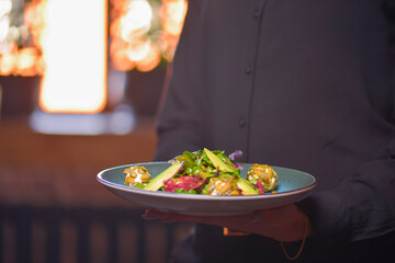 Waiter in black uniform, shirt, bringing the ordered healthy spinach and arugula salad with avocados.