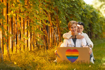 Portrait of two lesbians sitting on the grass with poster with LGBT sign and smiling at camera