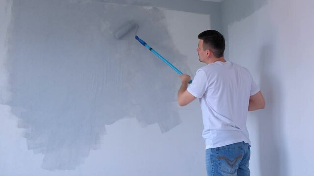 Man painter is painting wall in gray using paint rollers in new house himself. Young guy is making renovation in new apartment, flat, room. Construction and finishing work.