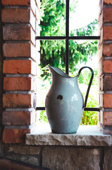 Stary metalowy dzban na wodę na tle ogrodu, po bokach ceglasta ściana