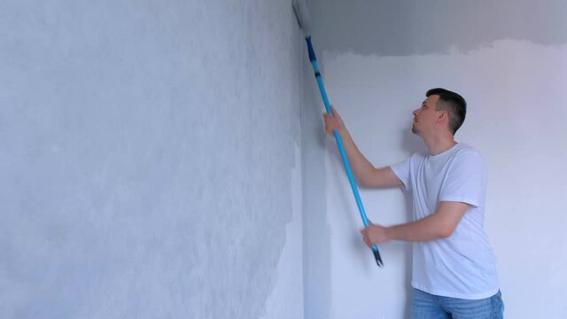 Man painter is painting wall in gray using paint rollers in new house himself. Young guy is making renovation in new apartment, flat, room. Construction and finishing work.