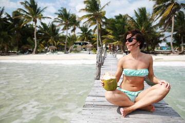 Brunette caucasian adult woman in swimsuit smiling with coconut on the wood pier on caribbean sea in Mexico