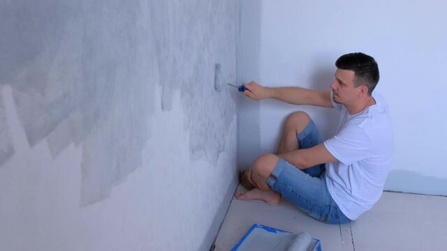 Portrait of young man painter is painting wall in gray using paint roller, side view. Builder guy is making renovation in flat room himrself. Construction and Finishing work at home, apartment.