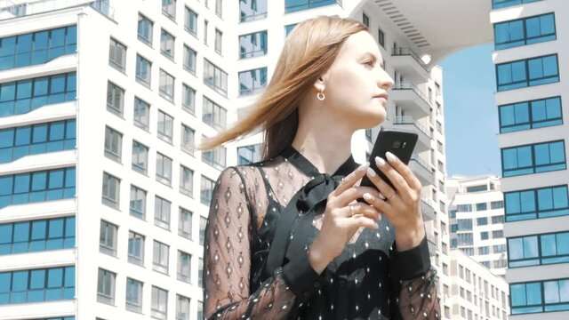 Confident happy attractive businesswoman female office  manager lady boss in black suit using phone looking at smartphone and waiting on the street outdoors near huge office buildings 