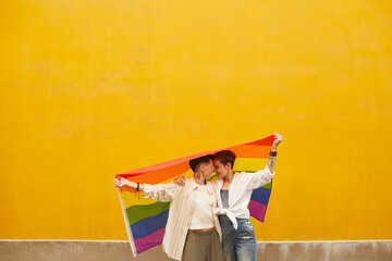 Two happy lesbians in love they standing against the yellow wall outdoors and holding colored flag - obrazy, fototapety, plakaty
