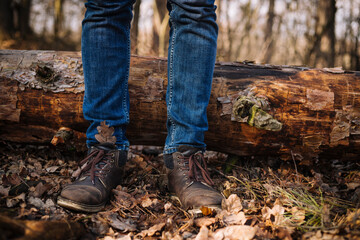 close up photo of human legs in old leather boots and blue jeans standing on the ground with dried leaves