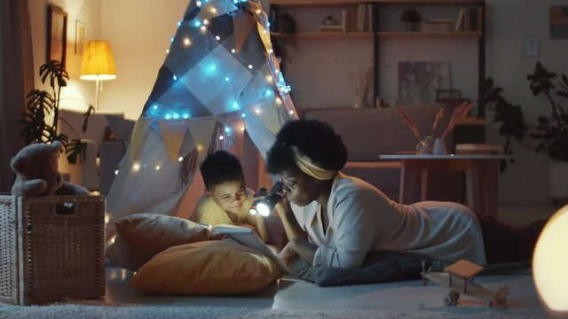 Little African American boy lying in teepee tent decorated with lights and listening to mom while she holding flashlight and reading fairytale to him in cozy dark room at home