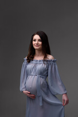 A young pregnant woman in a dress stands on a gray background.