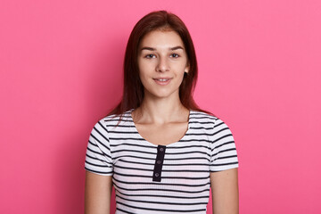 Fototapeta premium Young attractive teenage girl wearing striped casual t shirt, posing against pink background, looking directly at camera with charming smile, looks happy.