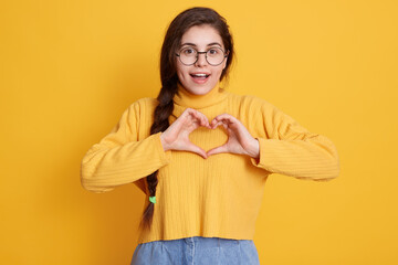 Excited young lady with pleasant smile showing heart gesture while posing against yellow background, girl with pigtail wearing sweater, expresses positive emotions.
