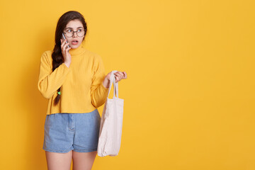 Portrait of shocked young girl with long dark hair posing isolated over yellow background with phone, talking to somebody with astonished facial expression.