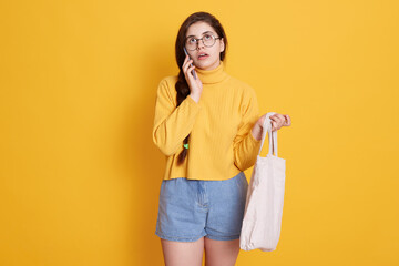 Young woman with cotton shopping bag talking on mobile phone while looking up, lady holding eco bag in hands, wearing yellow sweater and jeans short.