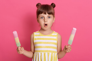 Girl with water ice creams in both hands wearing striped dress, having two knots, posing with astonished facial expression against pink background.