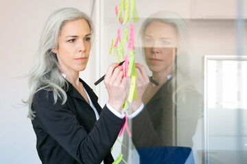 Content gray-haired Caucasian businesswoman writing on sticker with marker. Concentrated professional female manager sharing idea for project and making note. Brainstorming and business concept