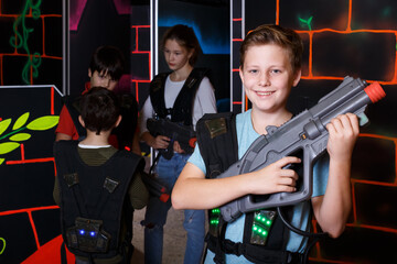 Portrait of laughing boy standing with laser gun on dark lasertag arena on background with his team